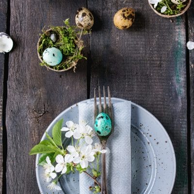 Nakrycie stołu kolorowe wielkanocne jajka przepiórcze z wiosennymi kwiatami wiśni, mech w doniczkach ogrodowych, puste talerze, sztućce na tle starego drewna. Ciemny styl rustykalny. Widok z góry z miejscem do kopiowania.